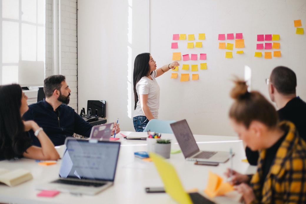 A woman pointing to Post-it Notes on a wall while people watch and listen